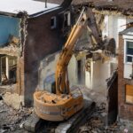 An excavator demolishes an old brick building at a construction site, creating debris and dust.