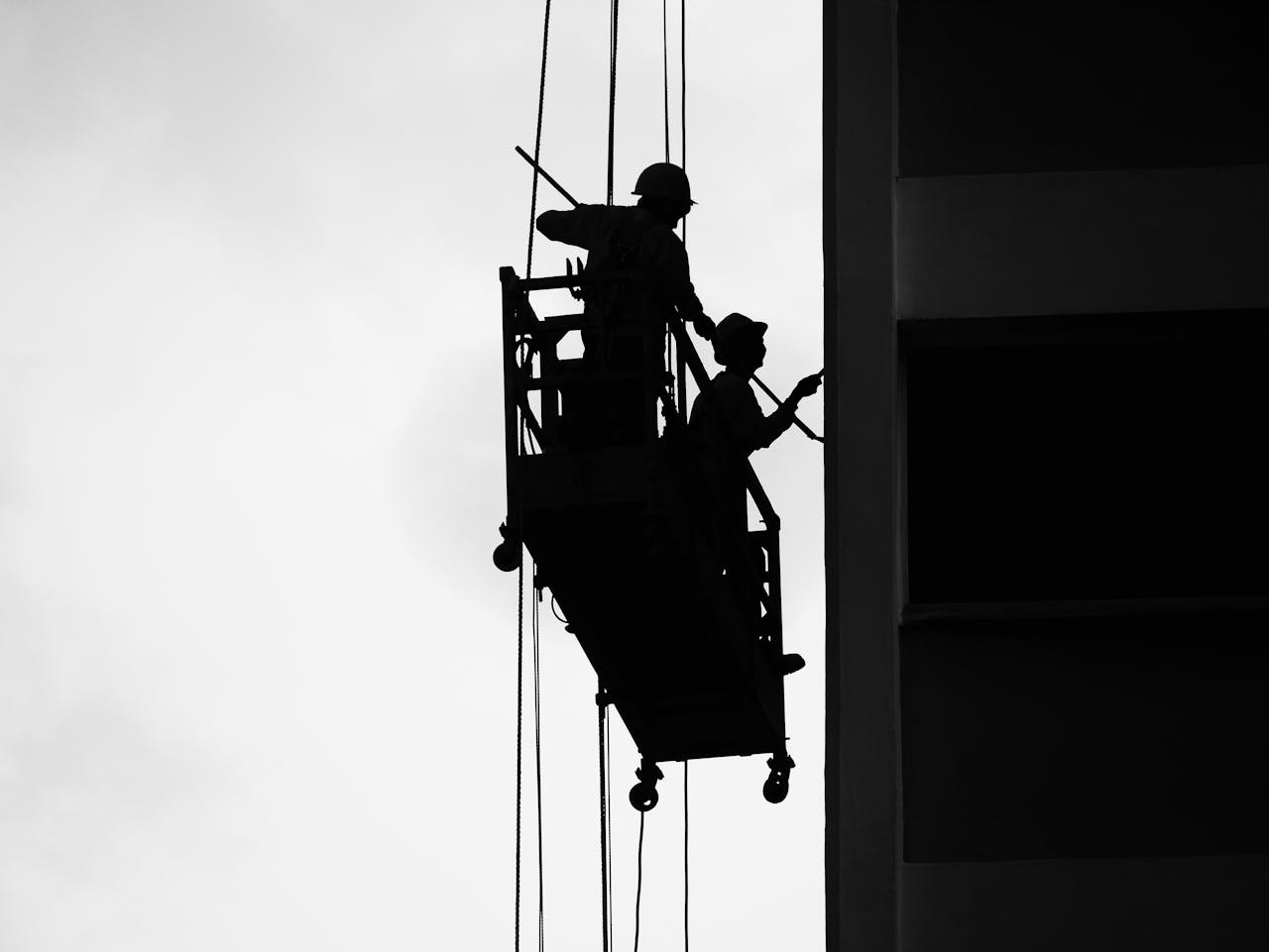 Two workers silhouetted on a suspended scaffold against a building facade.